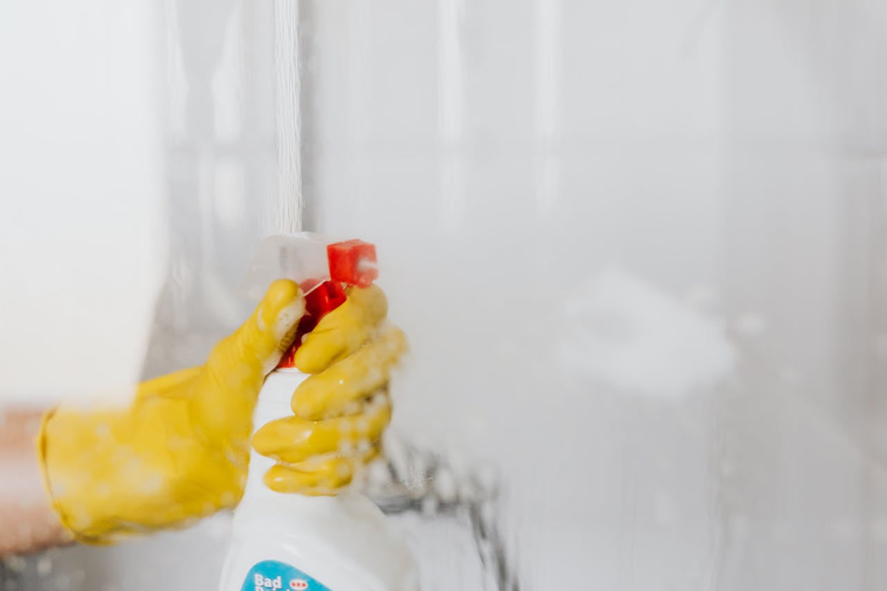services-02 Crop unrecognizable person in yellow latex protective glove spraying detergent from bottle with plastic dispenser at glass of shower cabin